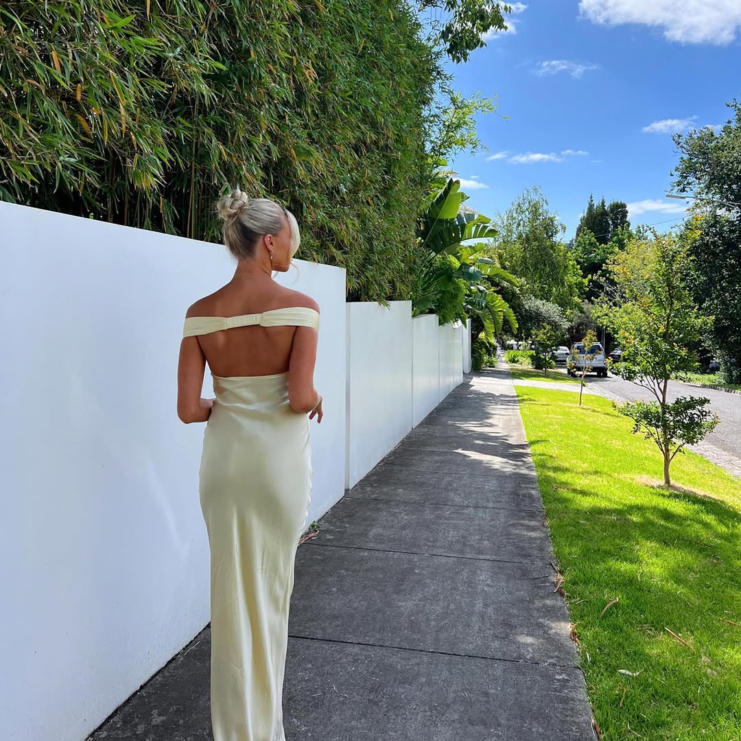 Woman in a white off-shoulder dress walking along a path with greenery and a white wall in the background.