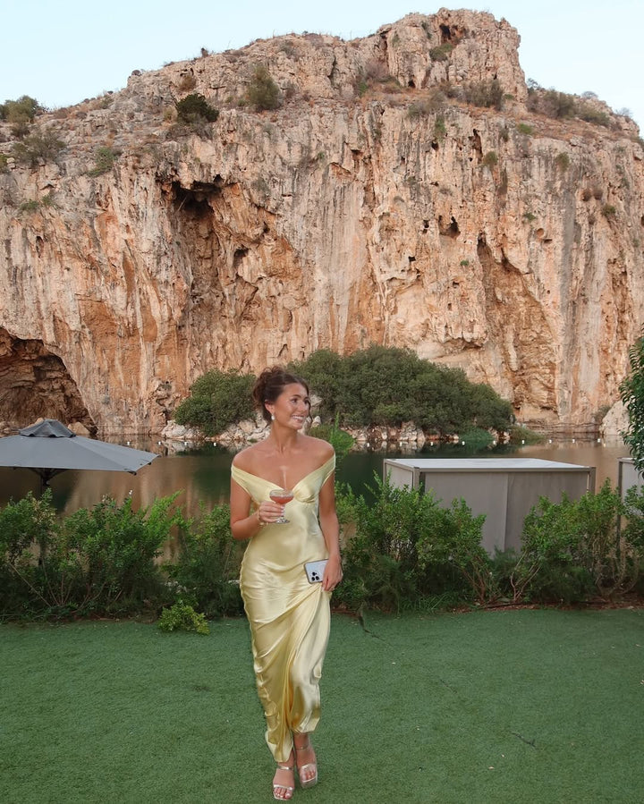 Woman in a yellow dress standing in front of a rocky cliff and body of water.