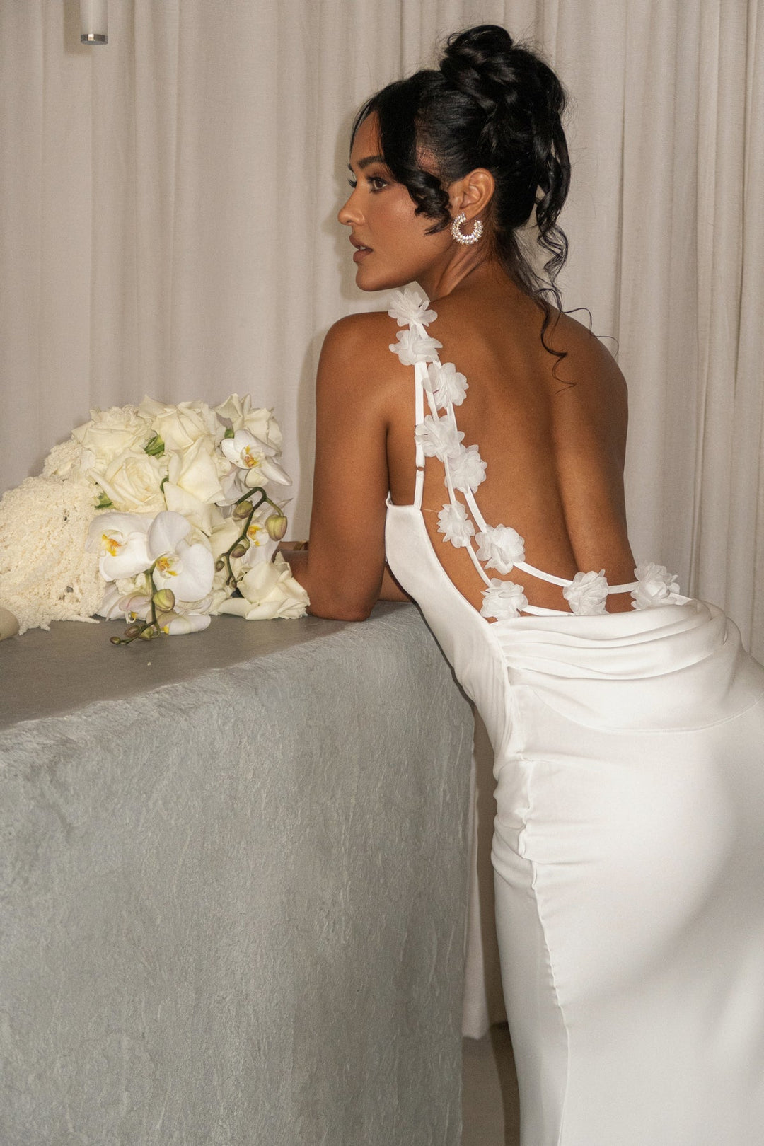 Woman in a white wedding dress with floral details, sitting next to a bouquet of flowers.