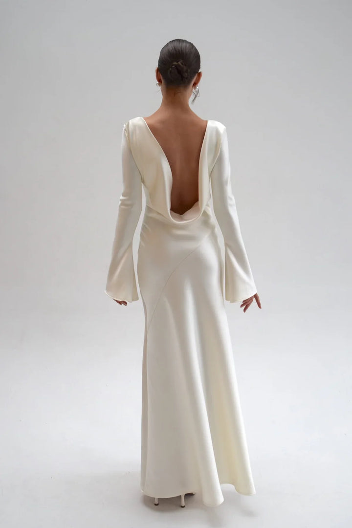 Woman wearing a long, elegant white dress with a deep backless design on a plain background
