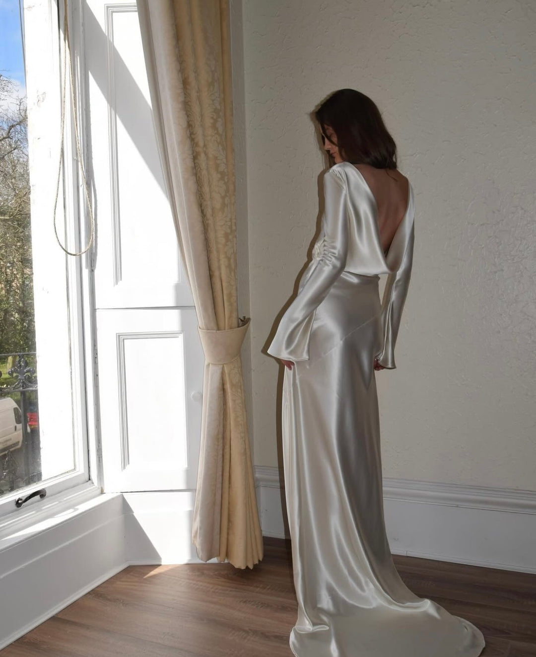 Woman in a white satin gown standing by a window with light streaming in.