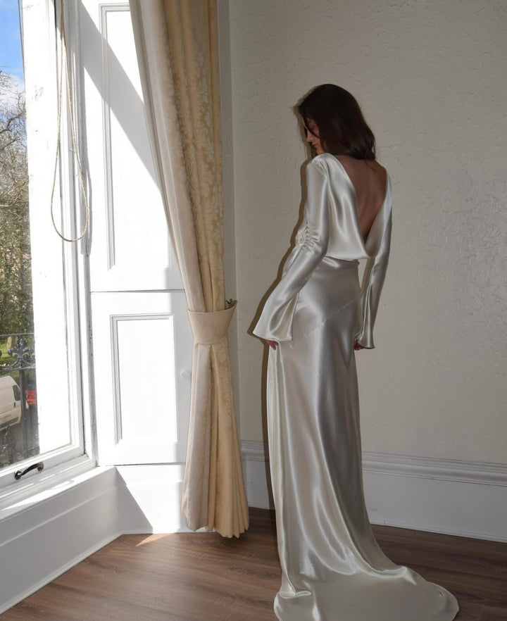 Woman in a white satin gown standing by a window with light streaming in.