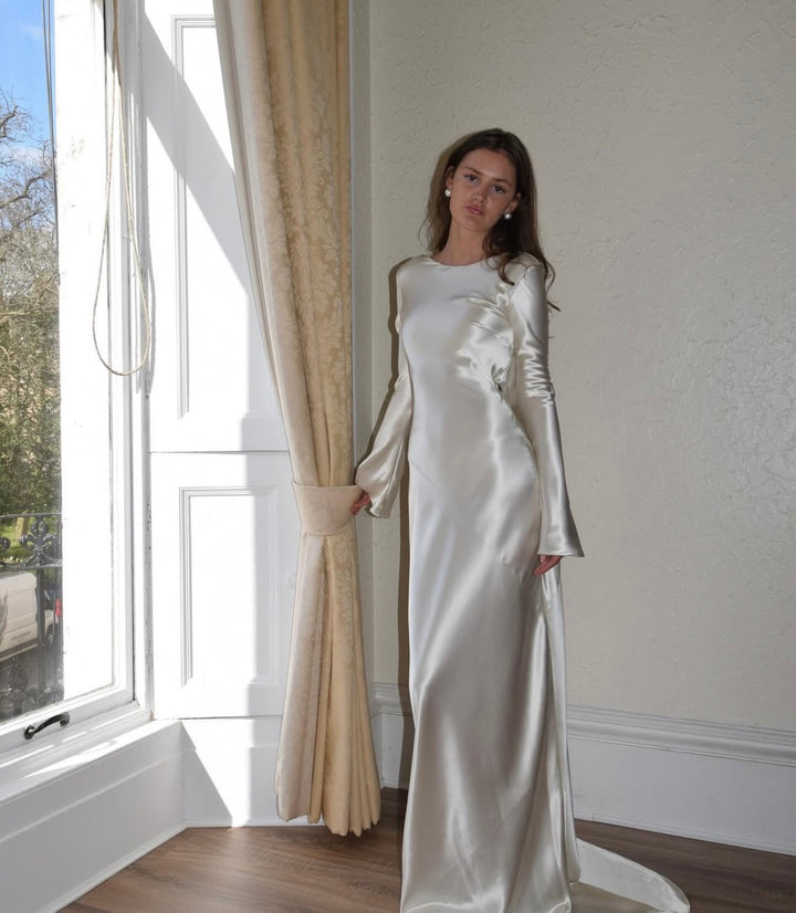Woman in a white satin dress standing by a window with beige curtains.