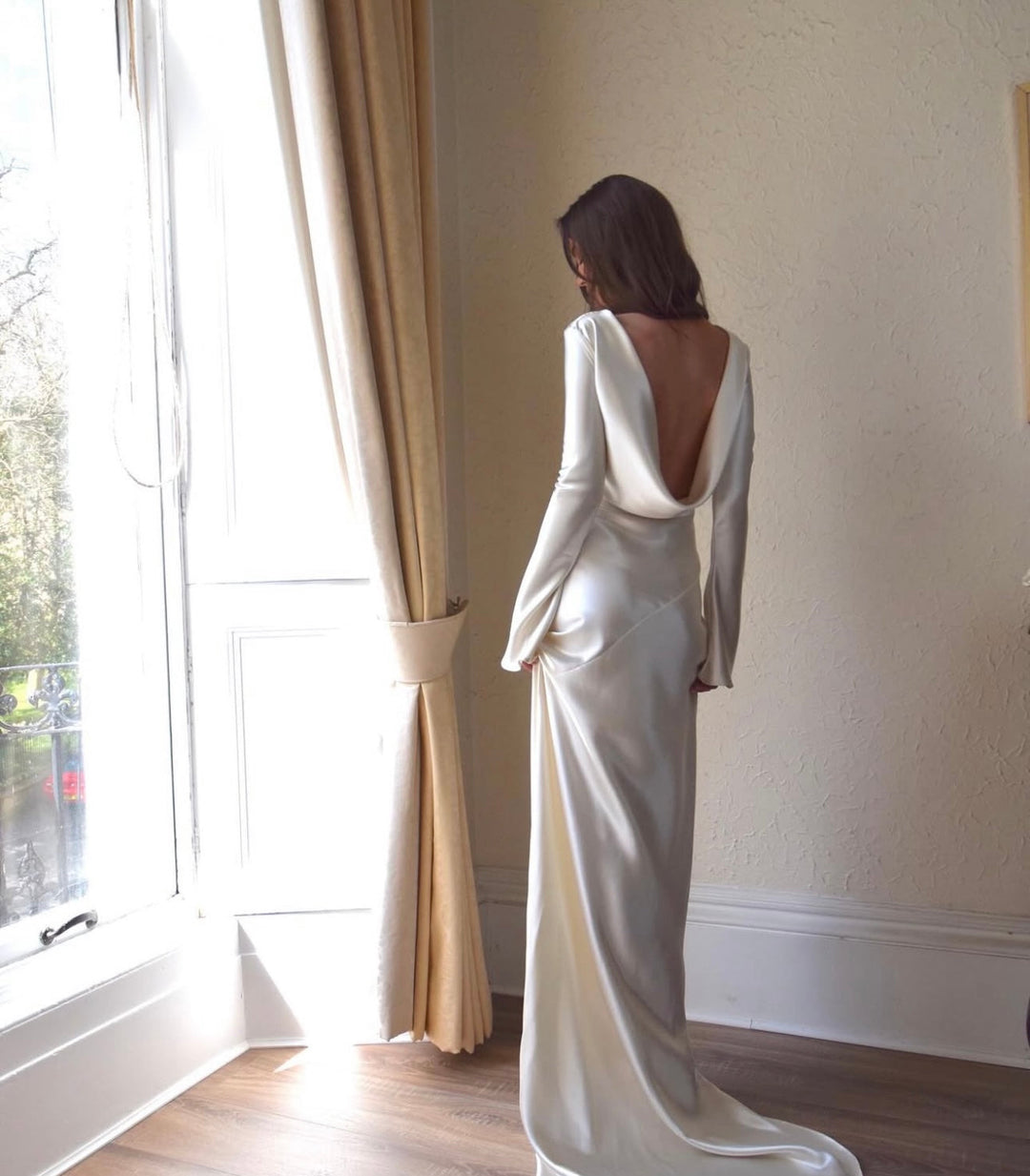 Woman in a white evening gown standing by a window with beige curtains.