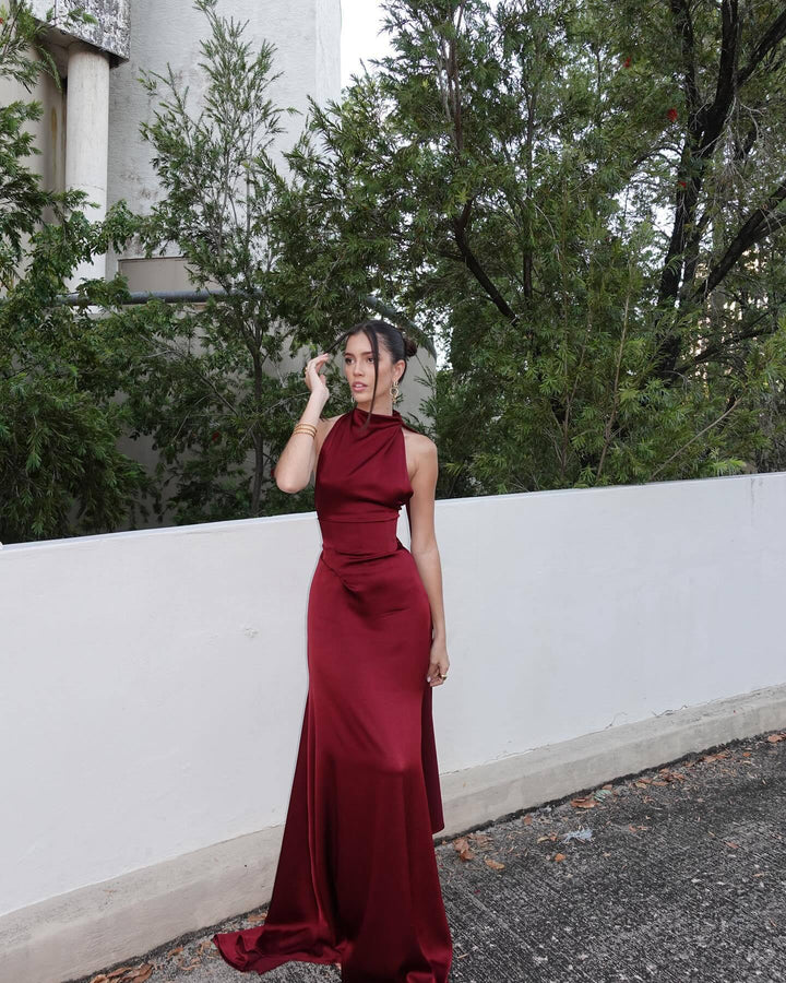 Woman in a burgundy evening gown standing outdoors with trees and a white wall in the background