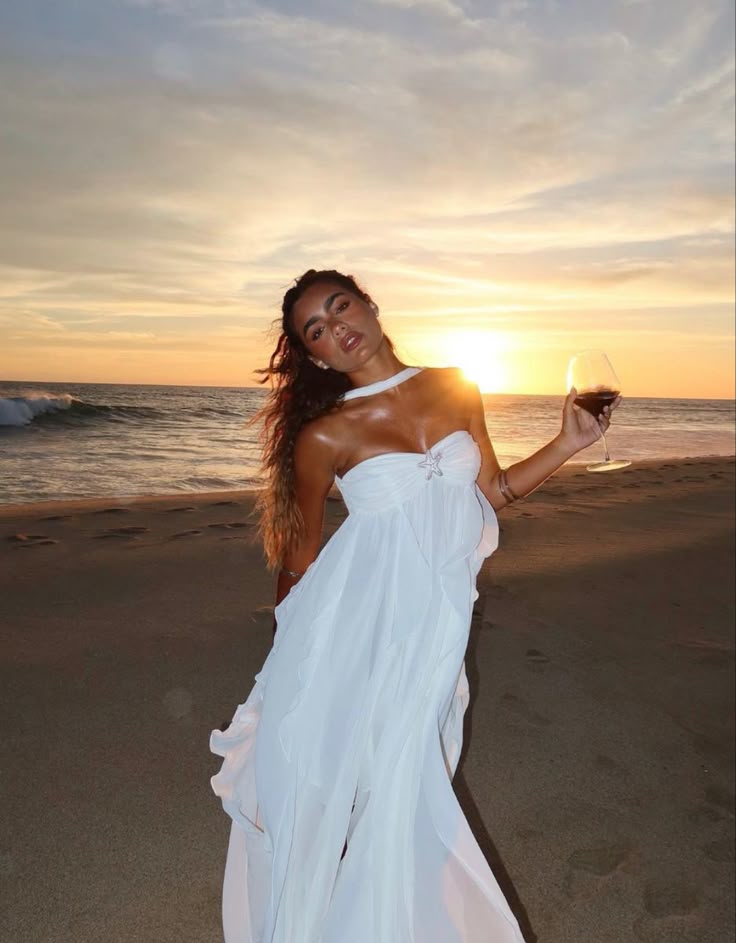 Woman in a white dress holding a glass of wine on a beach at sunset