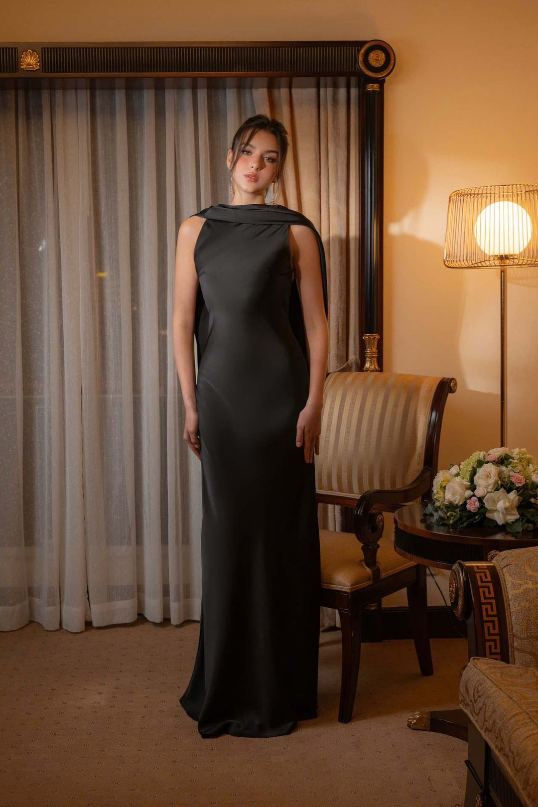 Woman in a black evening gown standing in a room with curtains and a lamp.