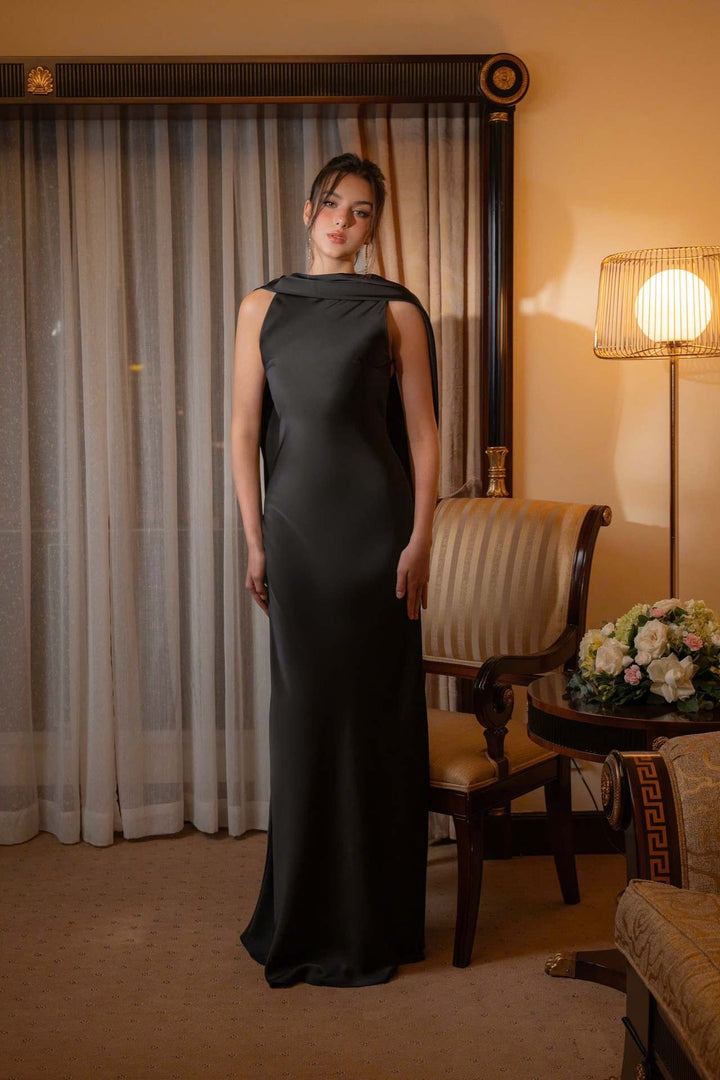 Woman in a black evening gown standing in a room with curtains and a lamp.