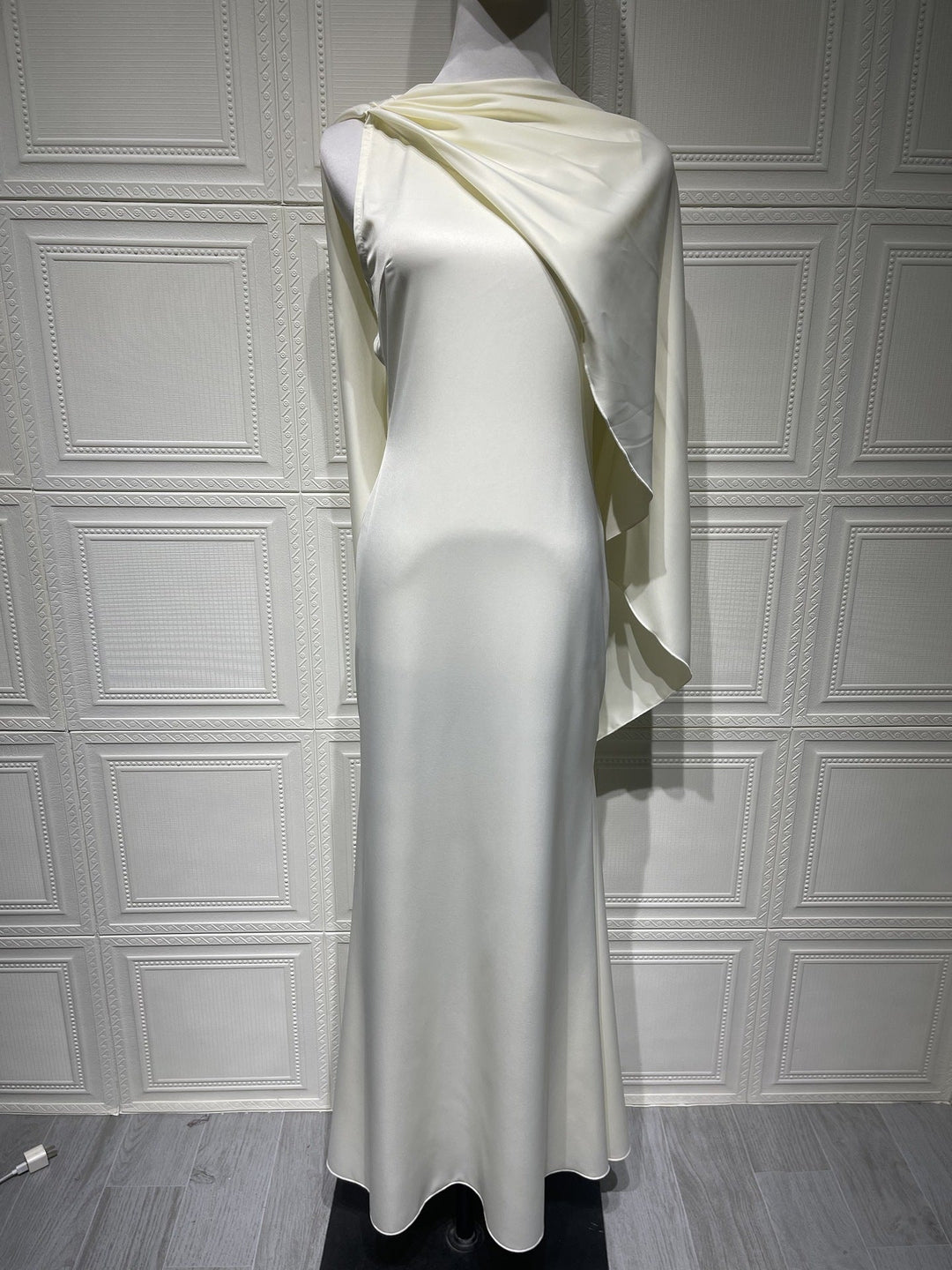 Mannequin wearing a long, light-colored dress with a draped fabric over one shoulder against a paneled wall.