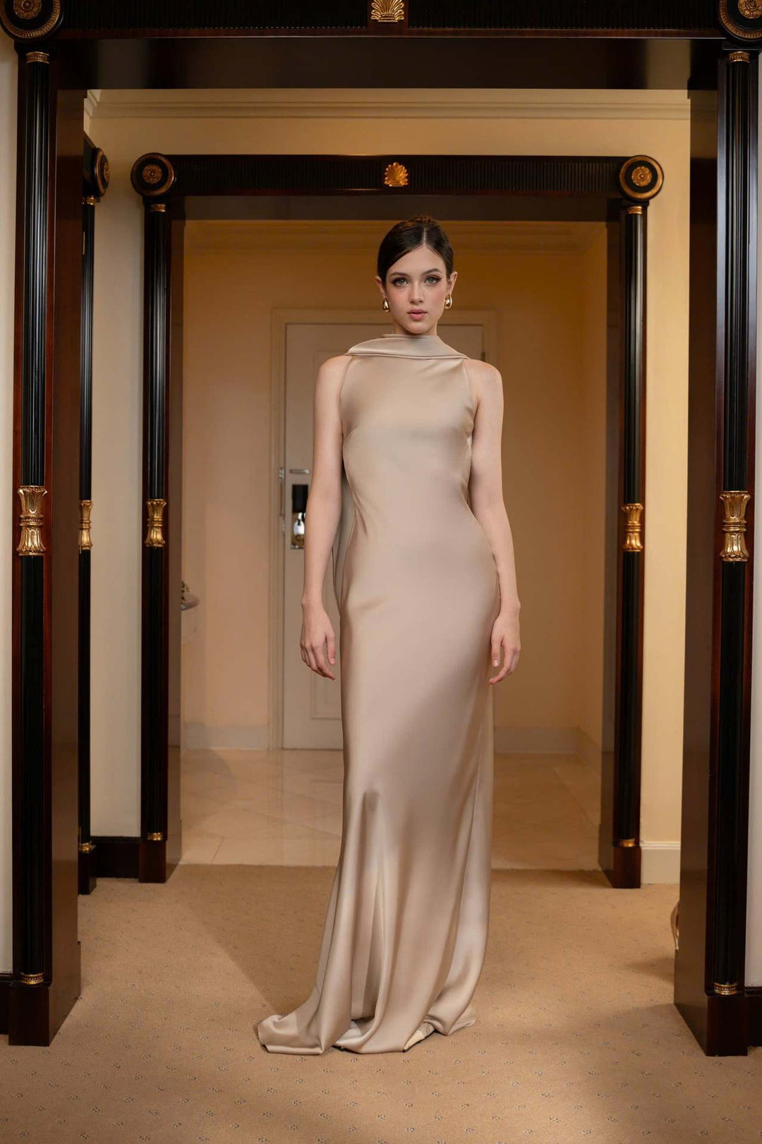 Woman in a beige evening gown standing in a hallway with decorative columns.