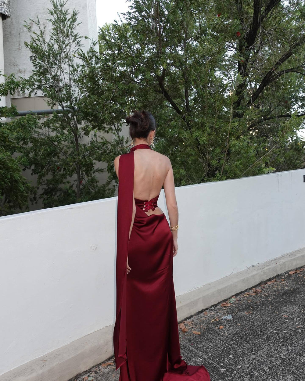 Person wearing a burgundy evening gown with a halter neck design, standing outdoors against a white wall and green trees.
