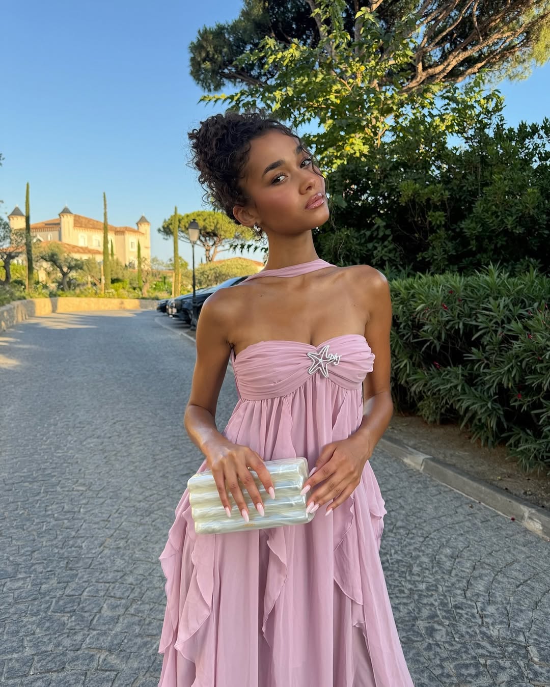 Woman in a pink dress holding a clutch on a street with greenery