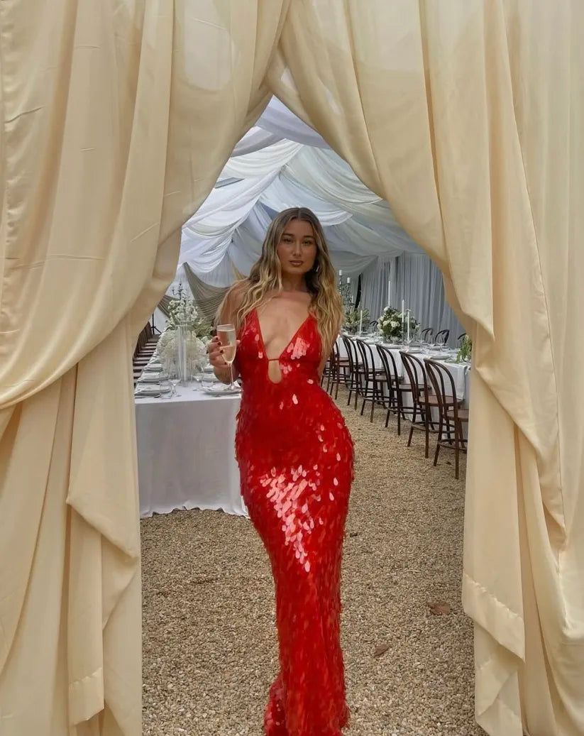 Woman in a red sequin dress standing between beige curtains with a decorated indoor setting in the background.
