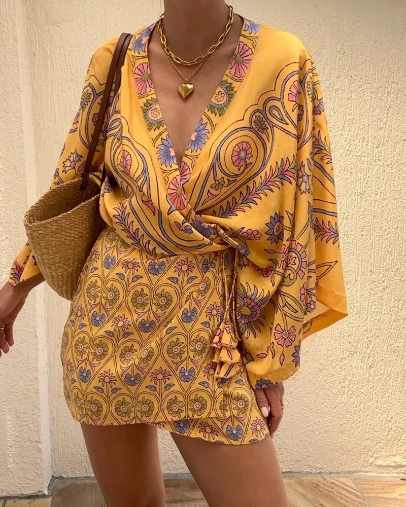 Woman wearing a yellow patterned dress holding a woven bag against a plain wall.