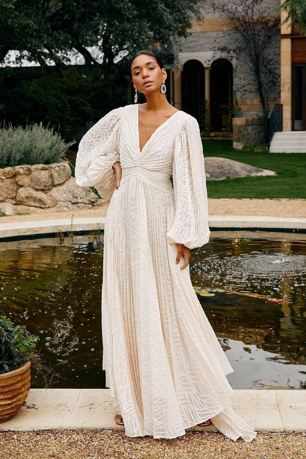 Woman in a white dress standing by a fountain with greenery and a building in the background