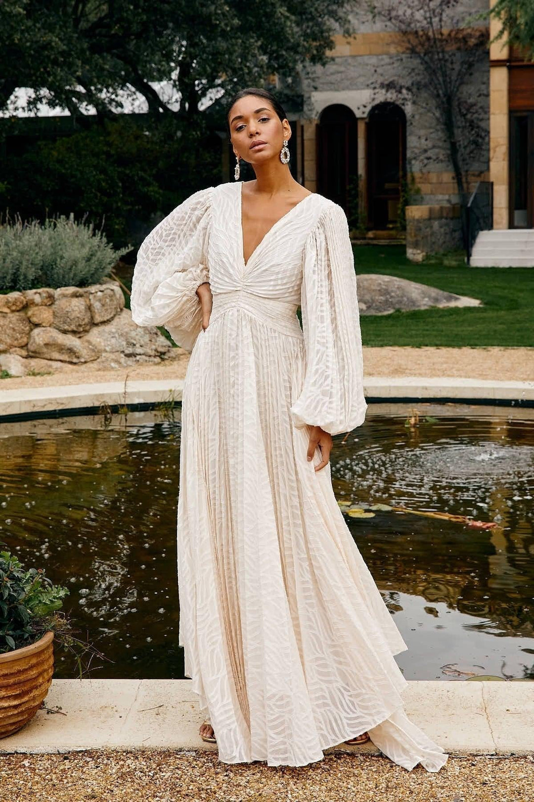 Woman in a white dress standing by a fountain with greenery and a building in the background