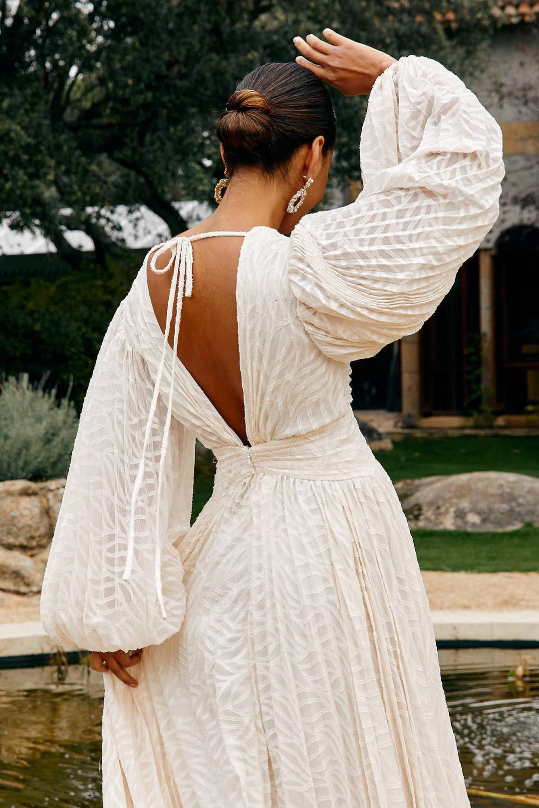 Woman in a white dress with long sleeves and open back standing by a fountain.